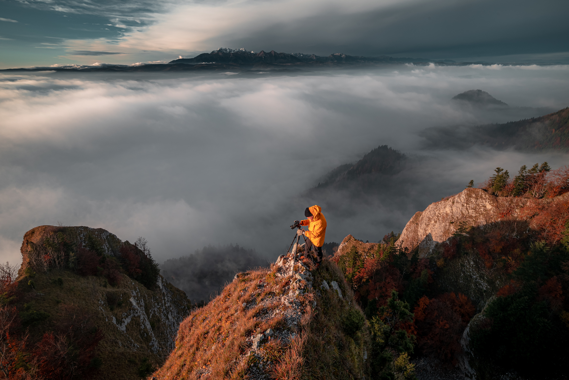Fotograf w żółtej kurtce robi zdjęcie na szczycie góry a obok unosi się mgła