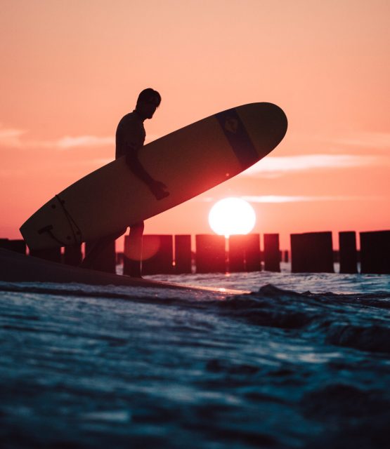 Surfer trzymający deskę pod pachą na tle zachodzącego słońca